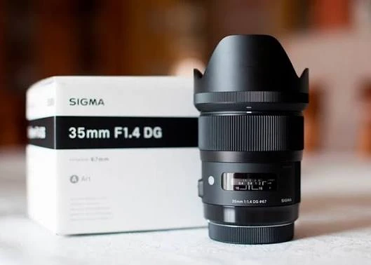 Об'єктив Sigma 35mm F1.4 DG Art чорного кольору з одягненою блендою стоїть поруч із білою фірмовою коробкою, на корпусі видно вікно шкали фокусування.